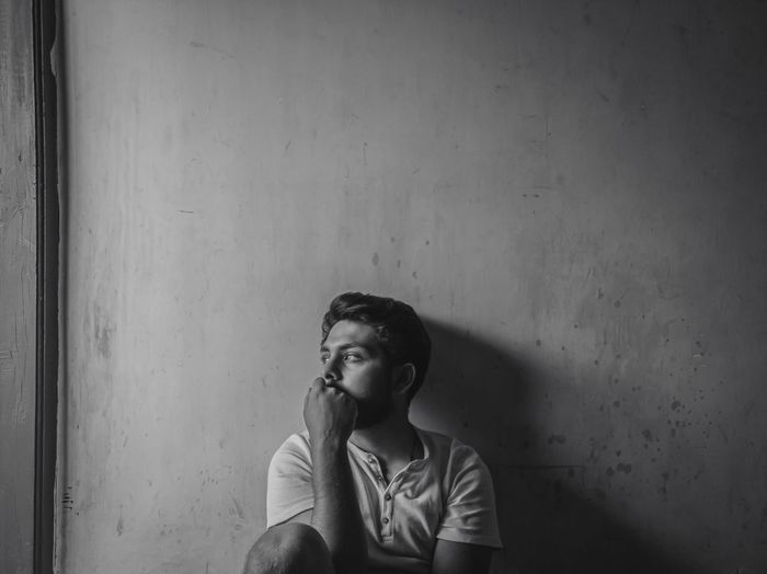 Thoughtful young man looking away sitting against wall