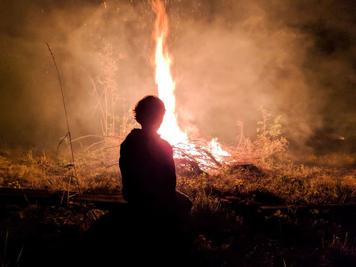 Silhouette boy sitting by campfire on field at night