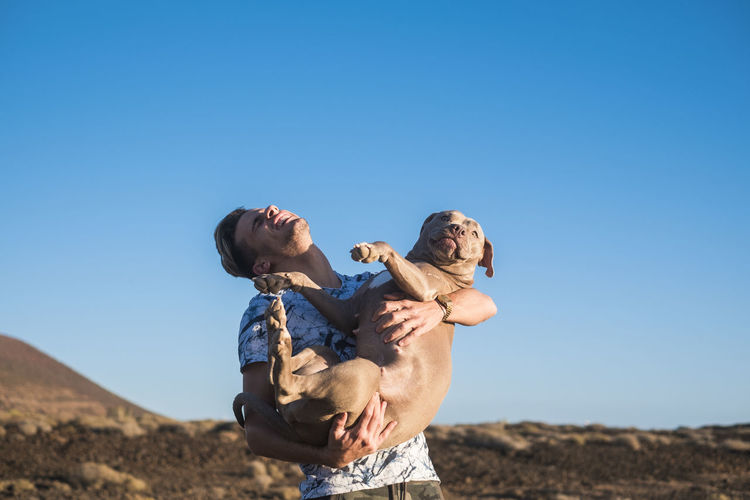 Cheerful young man carrying dog while standing against clear blue sky