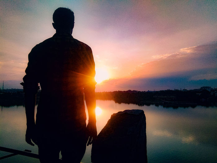 Rear view of silhouette man standing by lake against sky during sunset