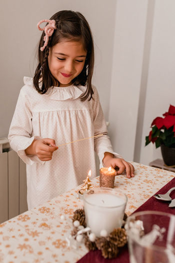 Cute girl in dress standing near festive table and lightning candles for celebrating christmas holiday