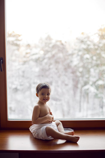 Baby boy in bath towel after washing sitting on the windowsill at the big window in winter