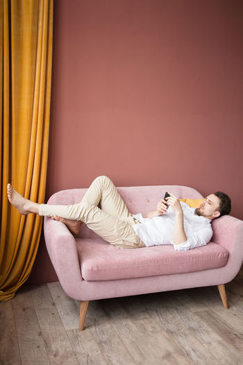 Young man in a white shirt works on his phone while lying on pink sofa