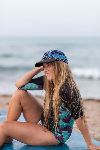 Side view of positive female surfer in swimwear and hat sitting on paddleboard on sandy shore against sea and looking away