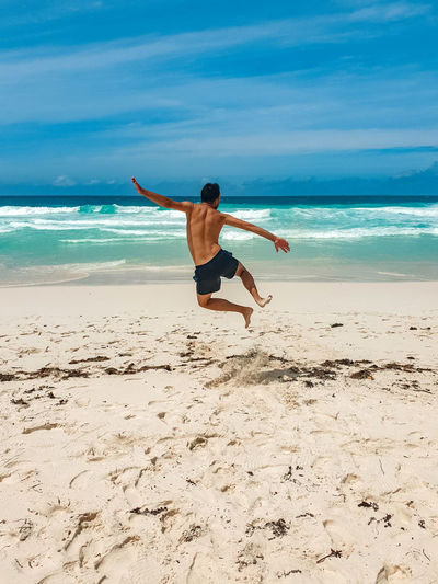 Rear view of shirtless young man jumping in air, clicking heels on tropical sandy beach