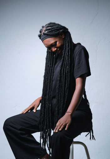 Smiling african american female with long black braids in casual clothes sitting on chair against gray background in studio
