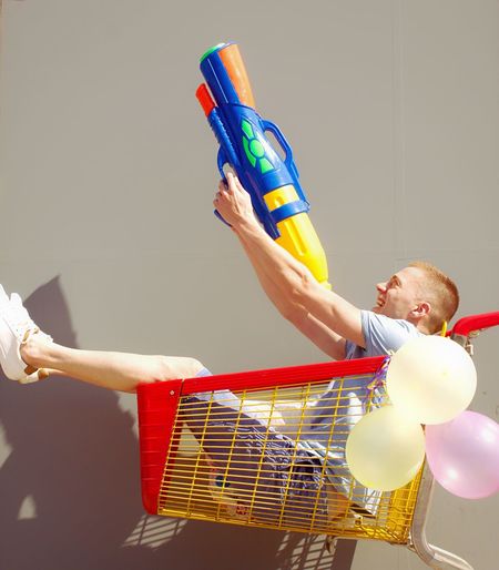 Man aiming with squirt gun in shopping cart against wall