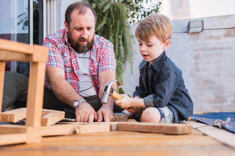 Bearded dad teaching son with hammer working with wood while sitting on boardwalk on weekend