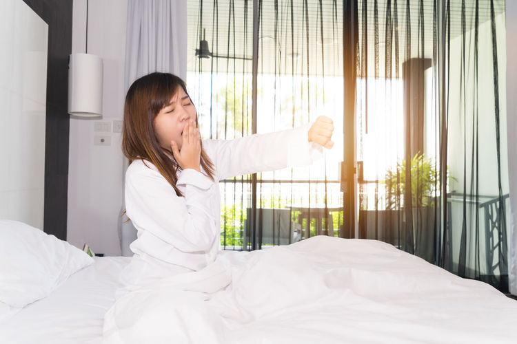 Young woman yawning while sitting on bed at home