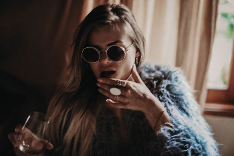 Young woman in sunglasses with glass of wine, blue fur coat and silver boho jewelry