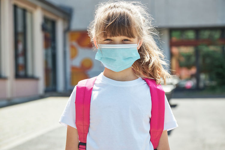 Close-up portrait of a girl in a protective mask and with a backpack in the yard of the school stot 