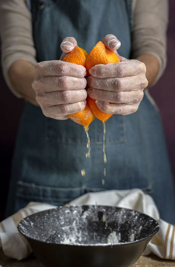 Crop hand of unrecognizable woman in apron squeezing fresh juicy cut orange over bowl while preparing dough at table
