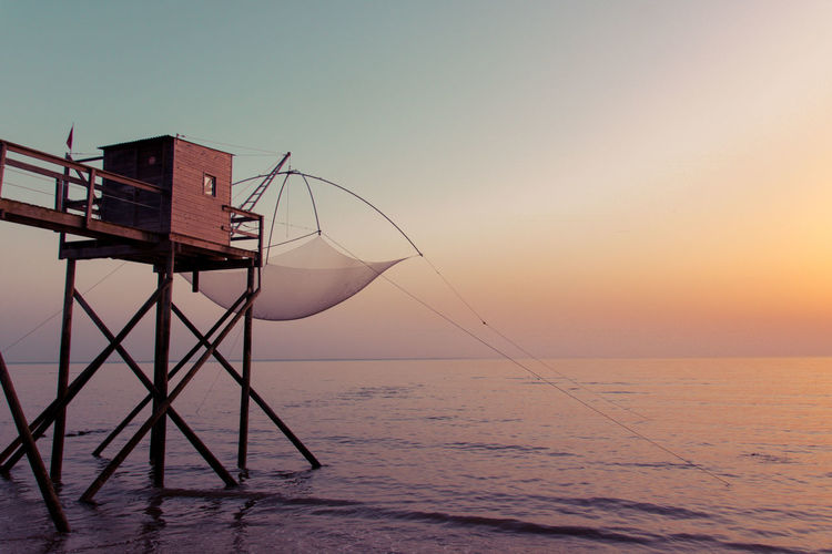 View of fishing equipment at stilt house over sea against sky during sunset