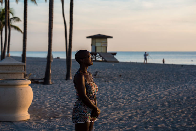 Thoughtful woman standing at beach against sky during sunset