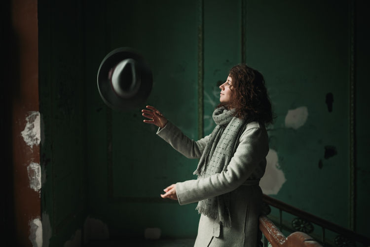 A young girl with wavy hair in a gray coat and scarf throws her gray hat up and smiles. Girl in an old house with green walls and stairs. Smile Green Color Hat Wall Coat Curly Hair Girl Gray Old Buildings Portrait Real People Scarf Stair Vintage Young Women