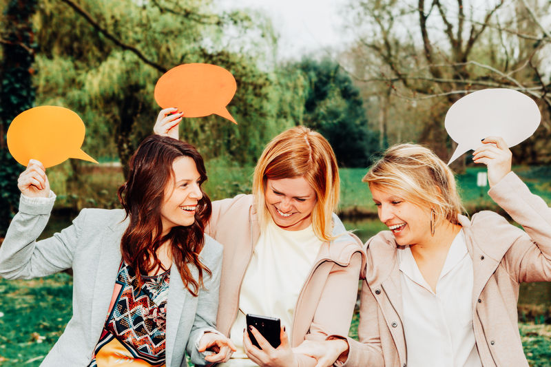 Women holding speech bubbles while using mobile phone against trees