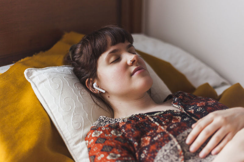 Young woman listening to guided meditation through wireless earphones while lying on bed