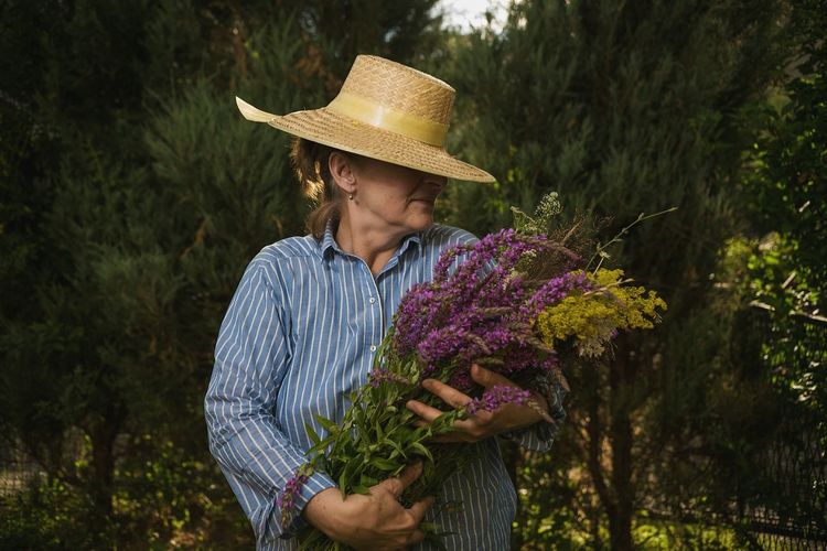 Mature female farmer holding fresh purple flowers against trees