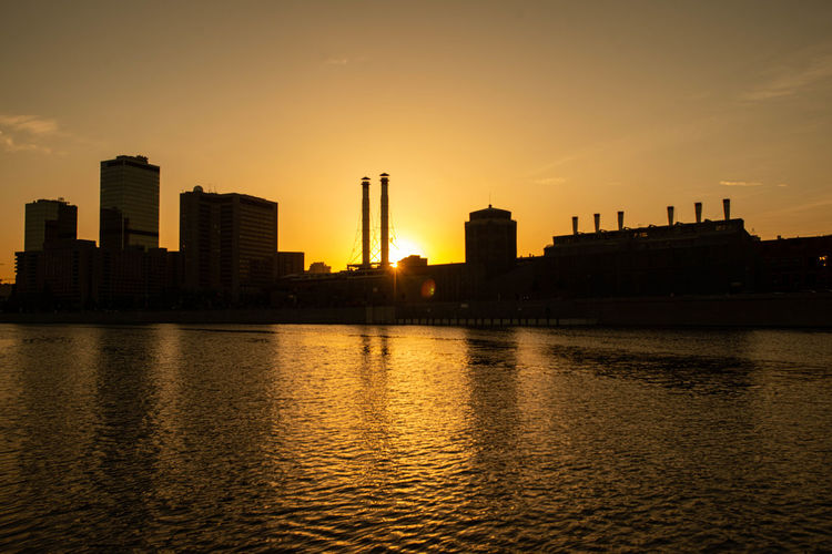 Silhouette buildings by river against sky during sunset