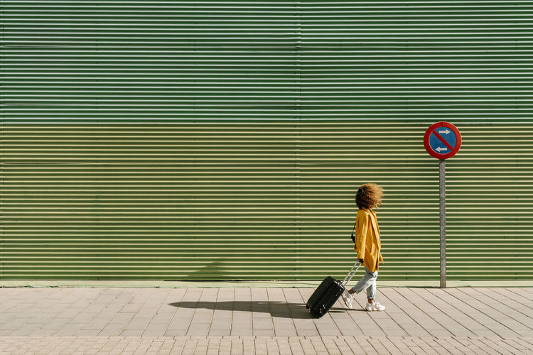 Afro woman pulling suitcase while walking on footpath by green wall
