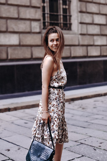 Young brunette woman in a long dress with black handbag smiling and posing on a street-style