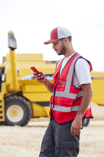 Side view of focused male worker in uniform texting message on cellphone while standing looking at screen near industrial combine harvester during work in agricultural field