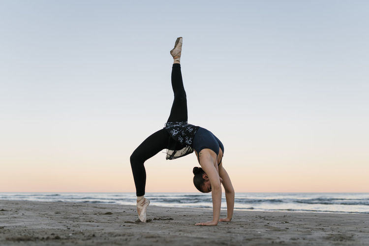 Flexible ballet dancer bending over backwards while practicing at beach against sky