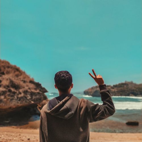 Rear view of young man gesturing while standing at beach against clear sky