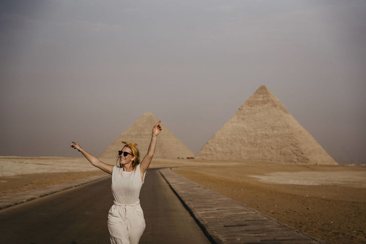 Female tourist standing with raised arms in middle of road leading to giza pyramids