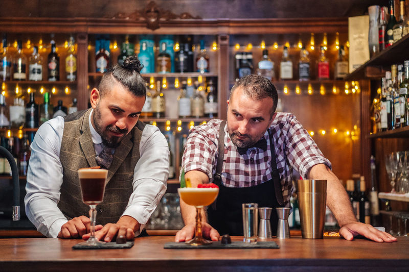 Bearded bartenders smiling while holding glasses of fresh cold sour cocktails at counter in bar