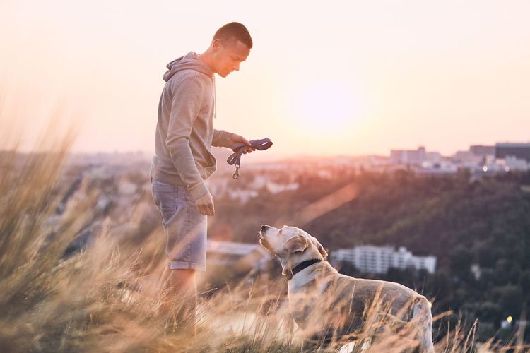 Side view of man with dog standing on field against sky during sunset