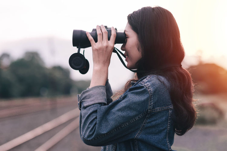 Woman looking through binoculars while standing against sky during sunset