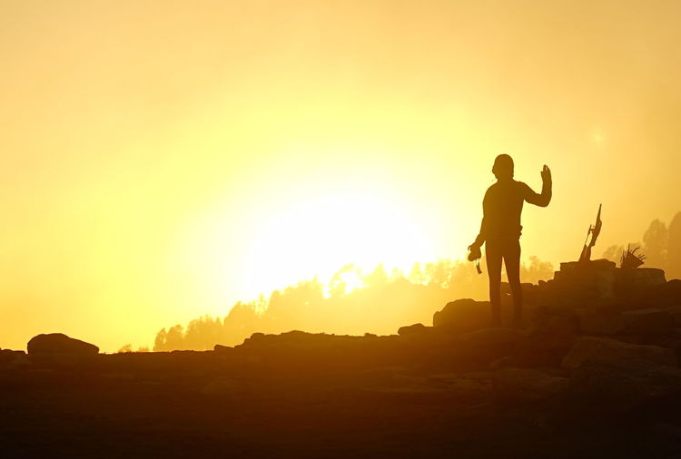 The silhouette of a woman who is waving to the camera, with the sunset behind her 
