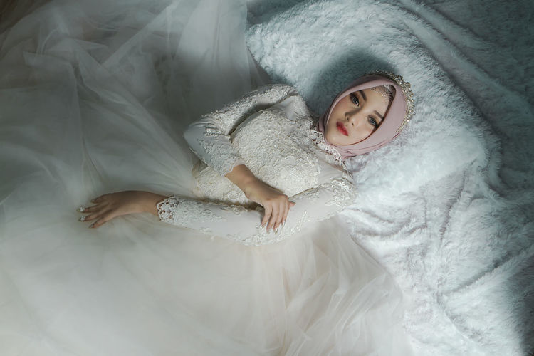 Asian muslim bride in white wedding gown with hijab or jilbab lying on bed. bridal wedding concept