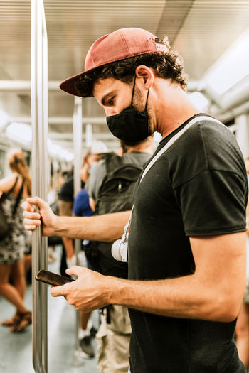 Side view of adult male passenger in casual outfit and black protective mask for coronavirus prevention browsing mobile phone during trip in subway train