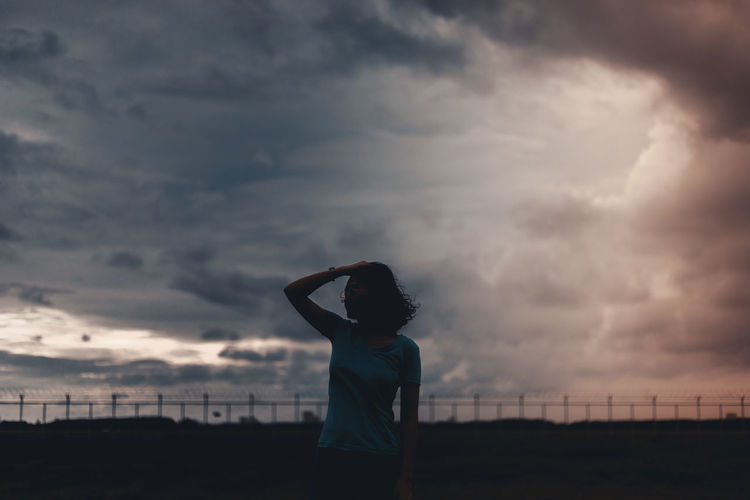 Silhouette young woman with hand in hair standing against cloudy sky during sunset