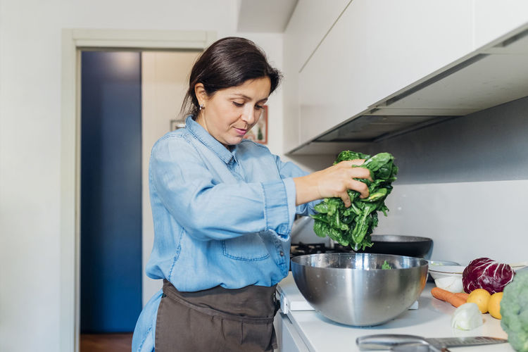 Woman taking out washed spinach from steel bowl in kitchen at home