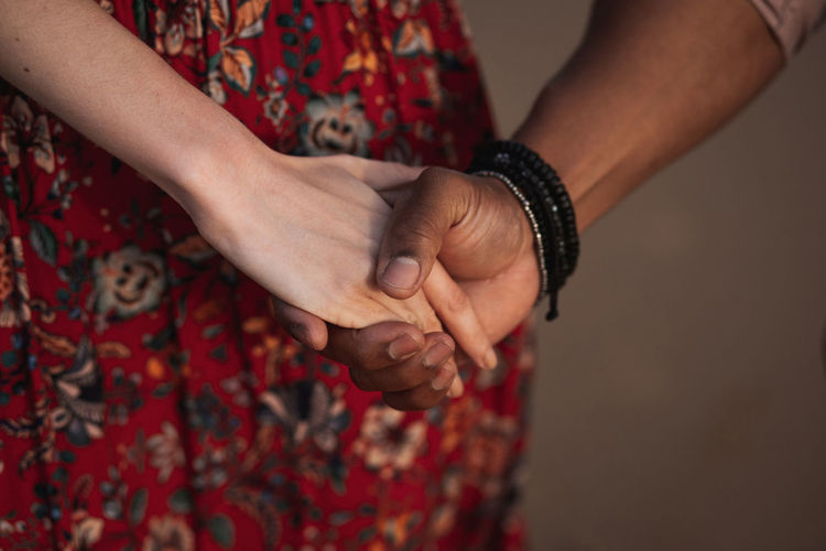 Crop anonymous woman in colorful dress and black man with bracelet on wrist holding hands while enjoying romantic moments together