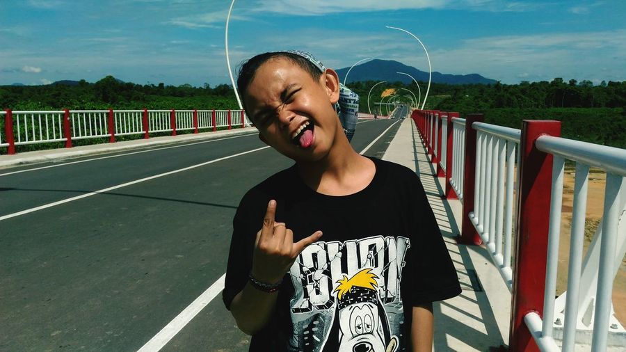 Playful boy gesturing horn sign while sticking out tongue on bridge during sunny day