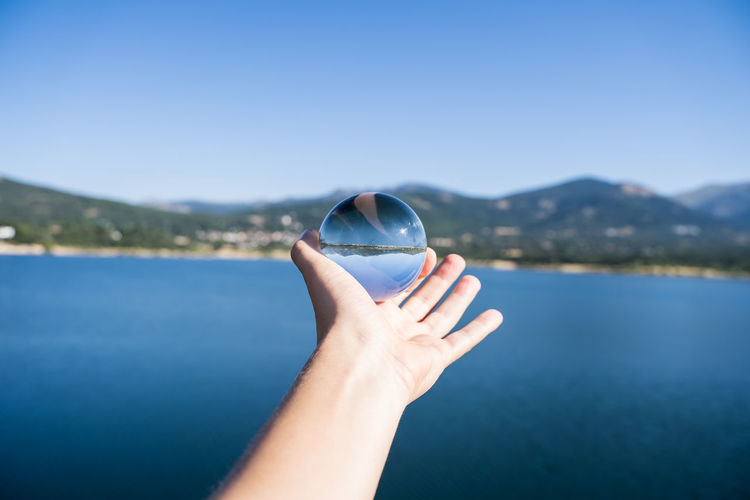Cropped image of hand holding crystal ball against lake