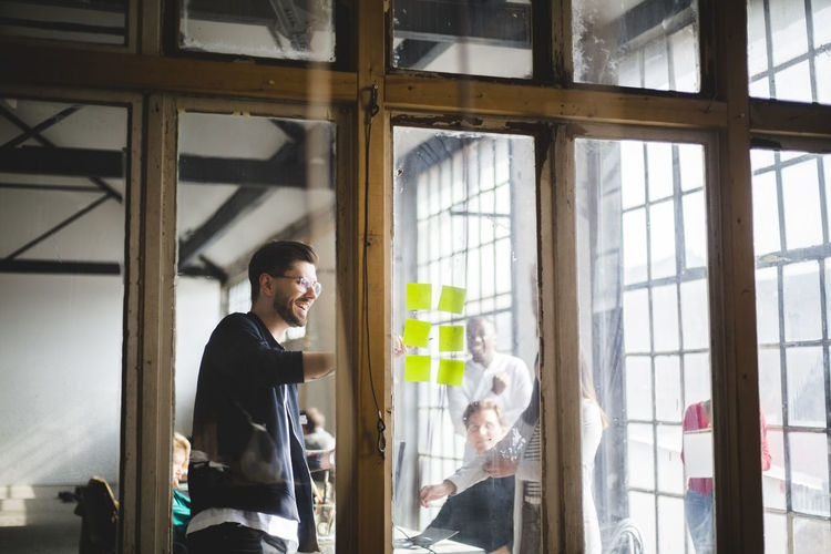 Smiling young it professional giving presentation to programmers while pointing at adhesive notes during meeting in offi