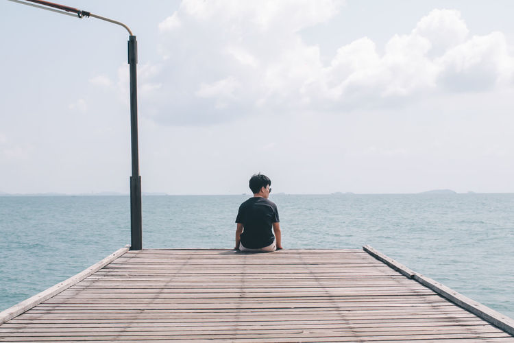 Rear view of man looking away while sitting on pier by sea