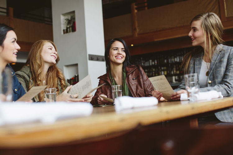 Happy female friends holding menu while talking at table in restaurant