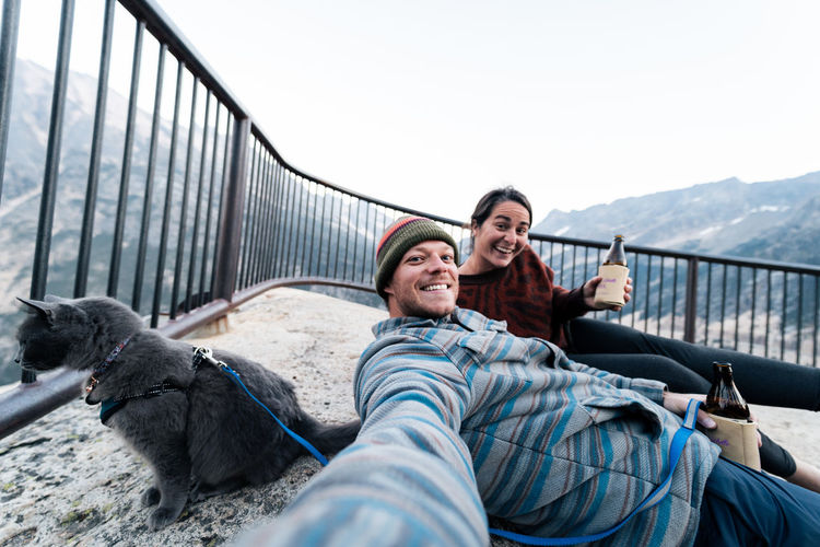 Young couple enjoys a time in wilderness taking selfies laughing and having fun with their gray cat