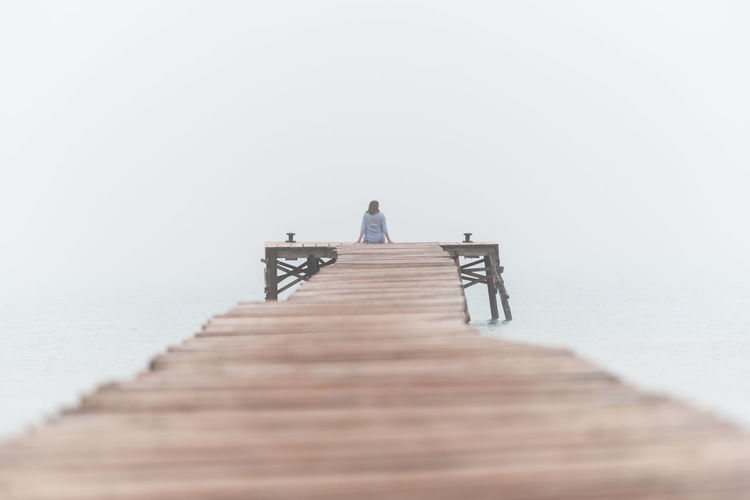 Back view of unrecognizable female sitting on wooden pier near sea on foggy morning in summer on playa de muro in spain