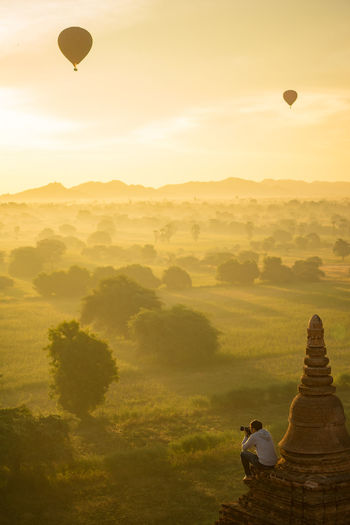 Side view of man photographing while sitting on temple against sky during sunset