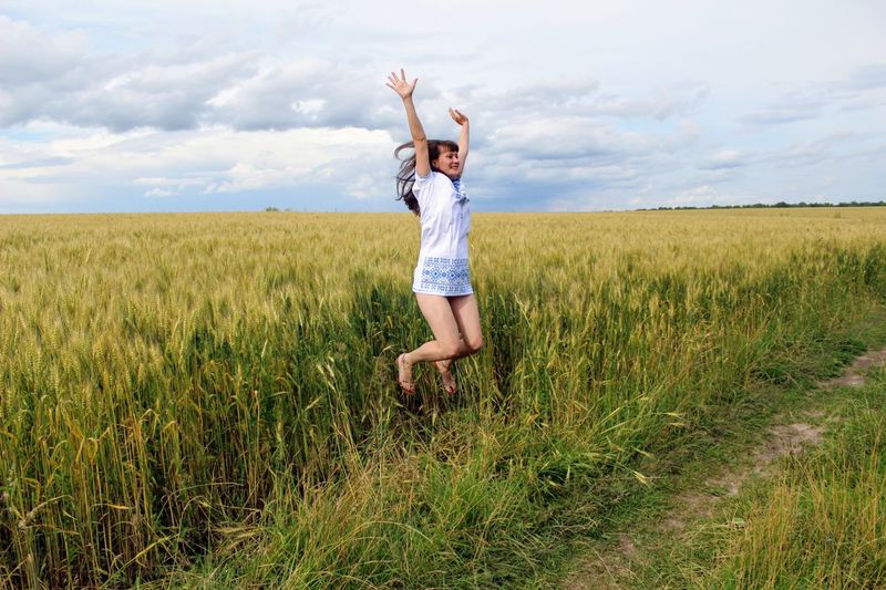 Cheerful young woman with arms raised jumping on field against cloudy sky