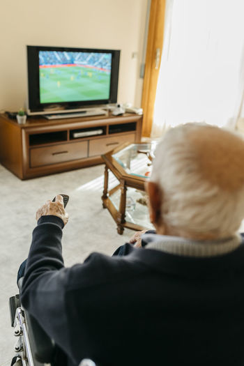 Back view of senior man watching tv at home using remote control