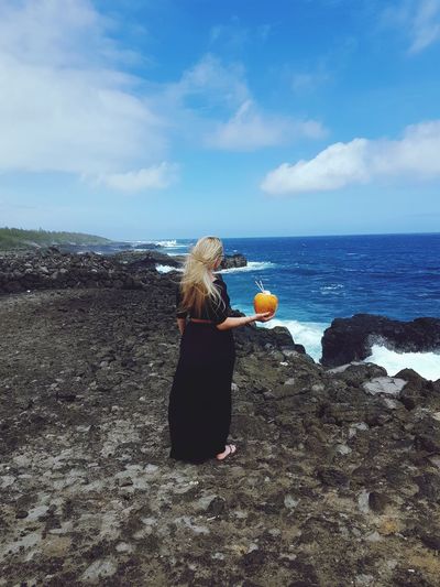 Woman holding coconut while standing on rocky shore against sky