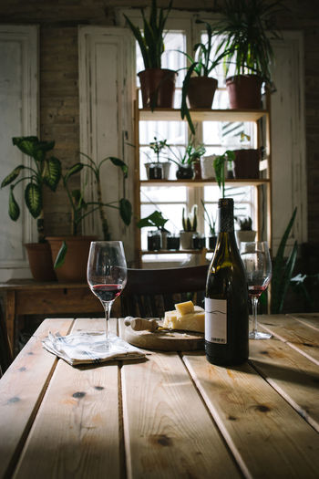 Open bottle and glasses with red wine placed near cheese on wooden table in rustic restaurant with potted green plants on window